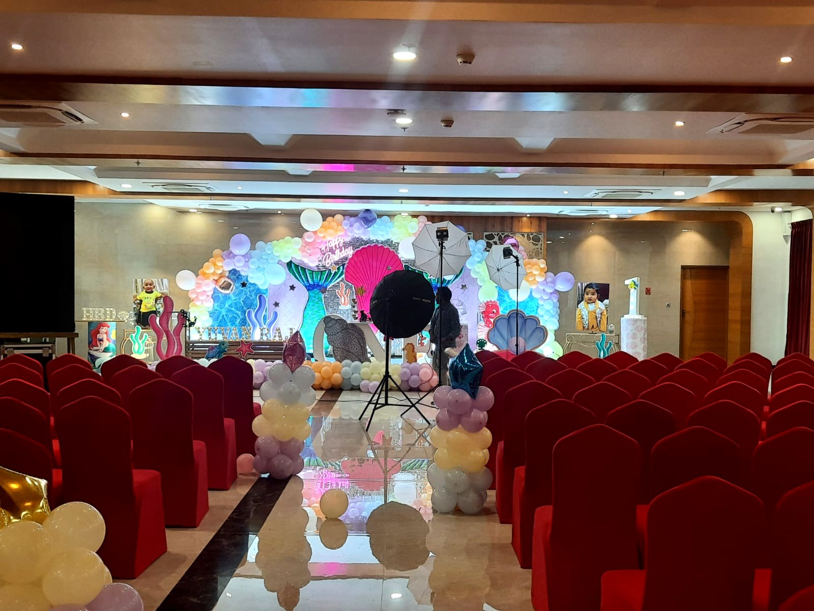 Birthday function hall with themed balloon backdrop and rows of red-covered chairs before guests arrive