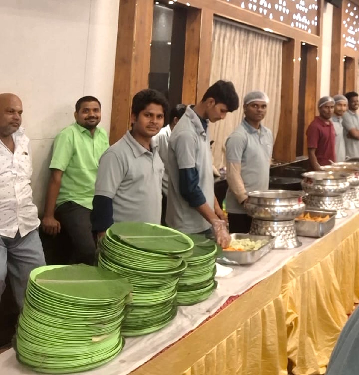 Sri Amrutha service team in uniform standing along a buffet line with stacked leaf plates and steel serving vessels