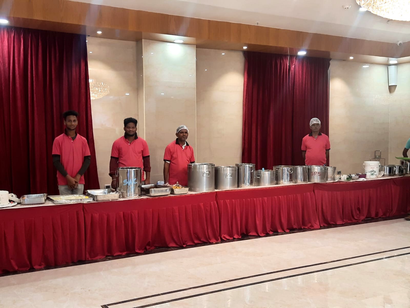 Sri Amrutha catering staff in maroon uniforms standing behind a red-draped buffet counter at a function hall